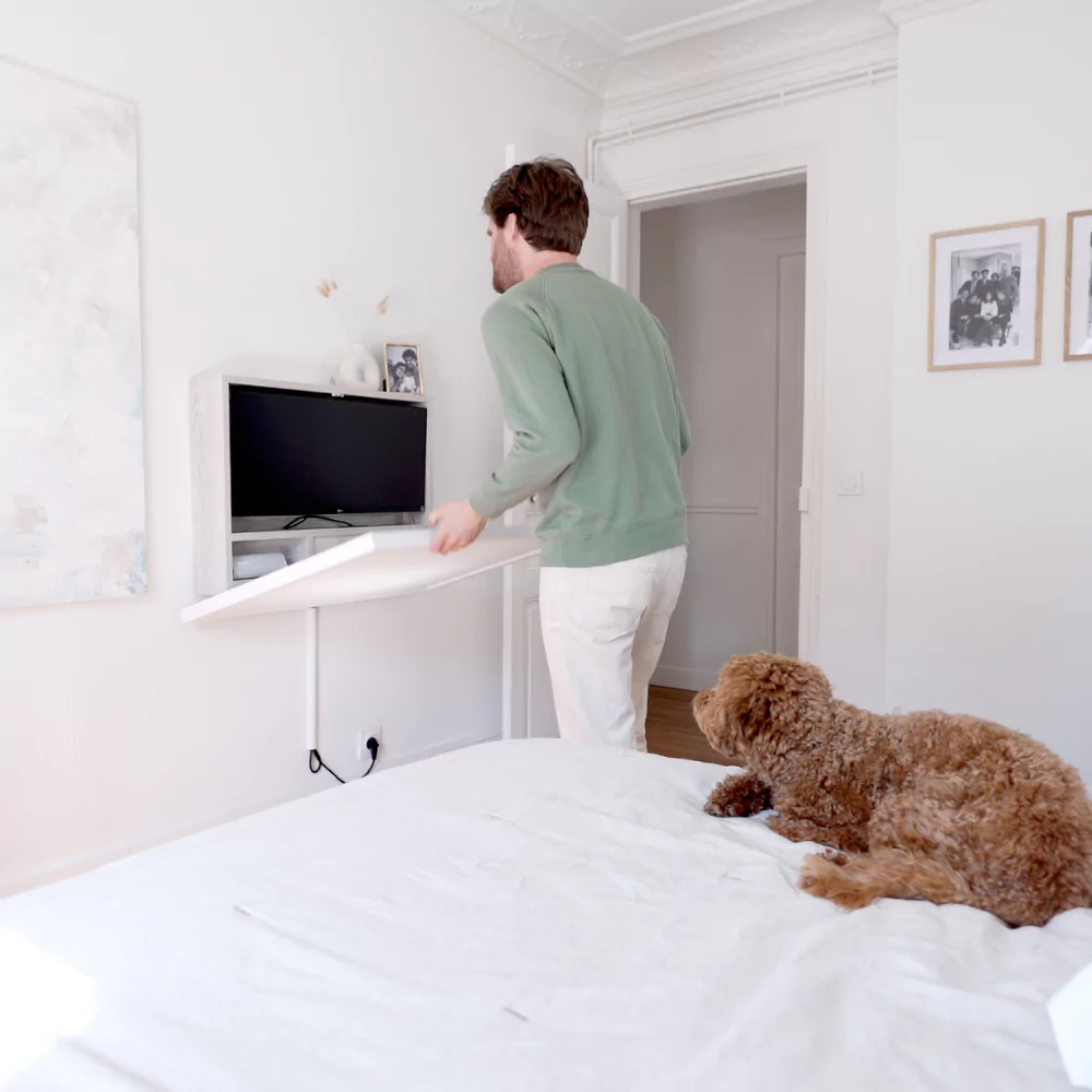 Tall man with green jumper closing his wall-mounted desk in a modern white interior and his #000 fluffy dog sitting on the bed behind him