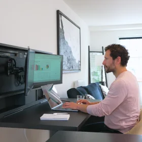 Person working at a wall-mounted fold-down desk with an integrated monitor setup and a laptop. The Murphy desk offers a compact, space-saving solution for a home office, blending into the living space for a seamless work-from-home experience.