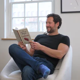 Person sitting in a comfortable chair near a window, smiling while reading a book titled ‘The Little Book of London.’ A relaxed setting that contrasts with a workspace, emphasising work-life balance.