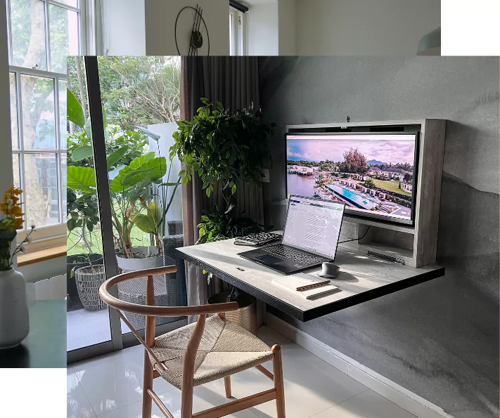 Minimalist home office with a wall mounted desk, integrated monitor, laptop and a wooden chair. Large windows and glass doors let in natural light, with greenery both inside and outside creating a calm atmosphere.